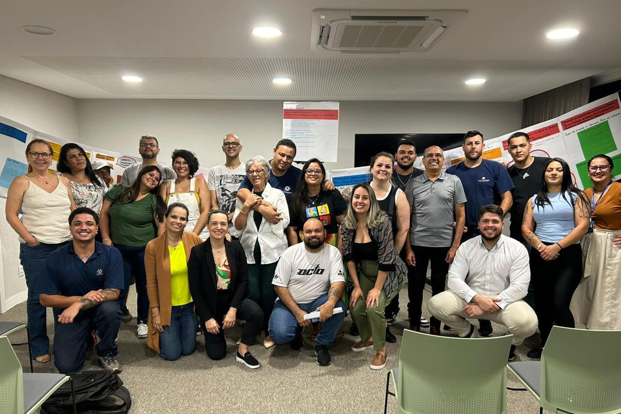 Participantes do grupo posam sorridentes em atividade da Escoop, com cartazes coloridos ao fundo, durante encontro de pesquisa e cooperação.