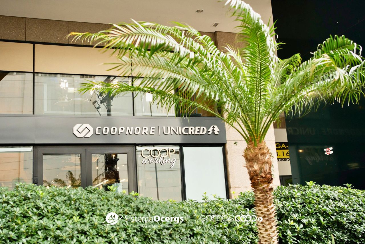 Descrição da imagem: Fachada da sede da Coopnore Unicred, com destaque para o letreiro da cooperativa e a inscrição "COOPworking" em uma das portas de vidro. Em primeiro plano, há uma palmeira e arbustos que compõem a paisagem externa.