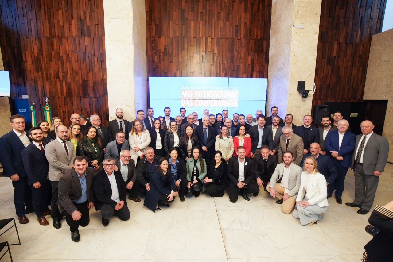 Grupo de dezenas de pessoas, entre homens e mulheres, posa para foto oficial em ambiente interno, diante de painel com os dizeres &quot;Ano Internacional das Cooperativas&quot;. O cenário sugere solenidade institucional ligada ao cooperativismo.