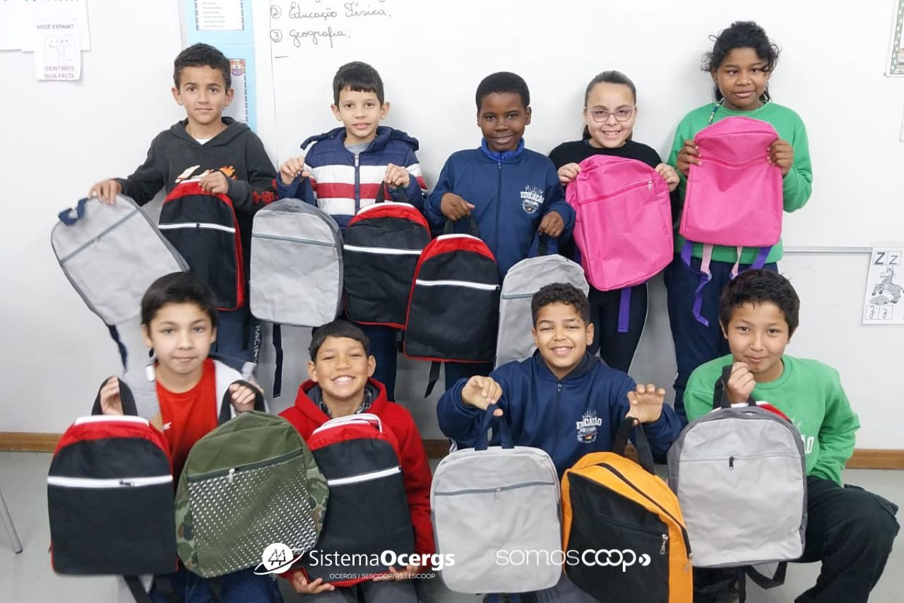 Alunos da Escola Estadual Fernandes Vieira, em Lajeado, posam sorridentes em sala de aula segurando mochilas coloridas doadas pela Unicred Premium em ação de educação financeira.