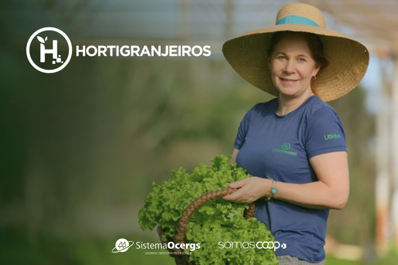Produtora rural sorri enquanto segura uma cesta de alface, usando camiseta e chapéu de palha. Ao fundo, o logo do Hortigranjeiros e as marcas do Sistema Ocergs e SomosCoop.
