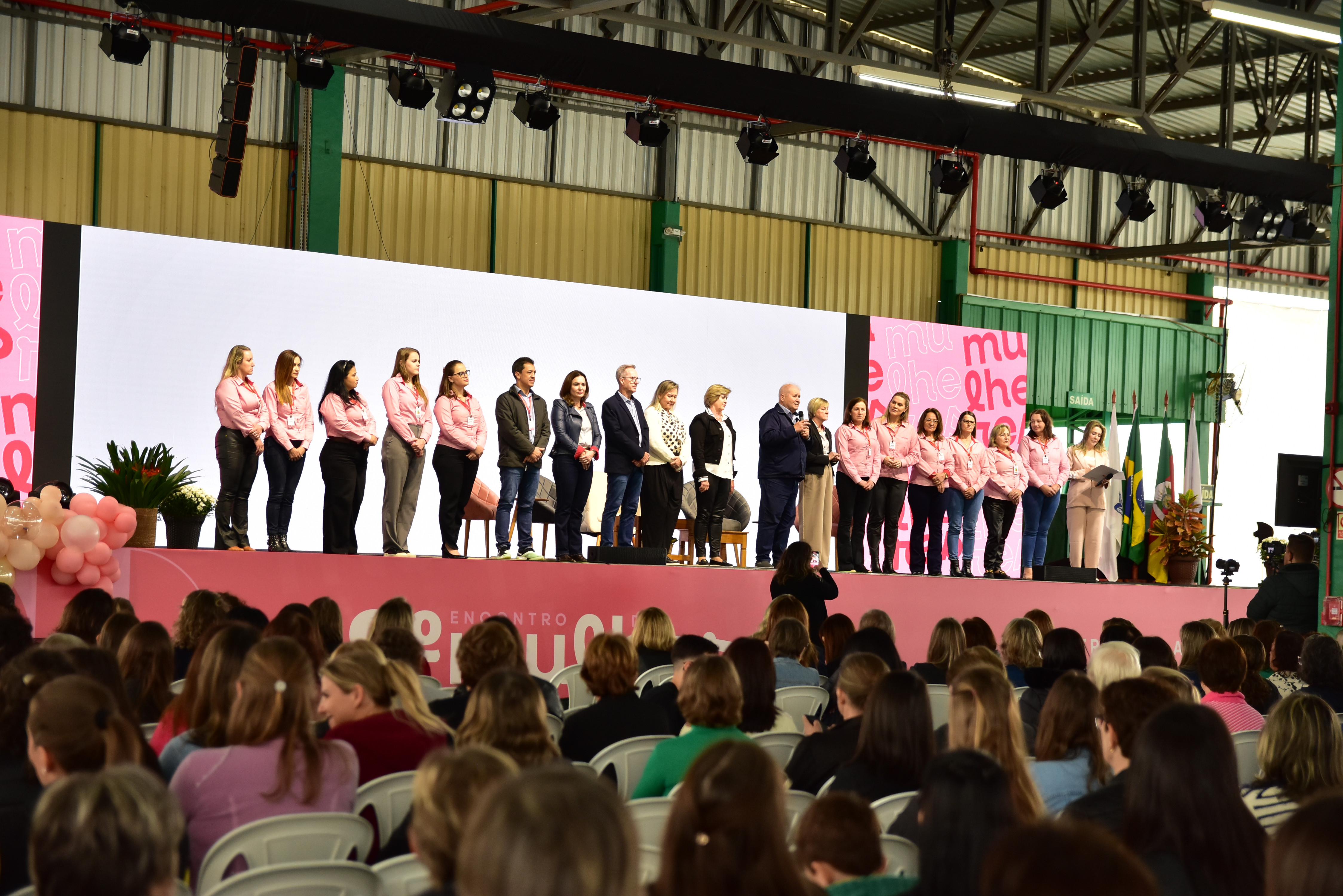 Imagem de um evento com uma grande plateia e um grupo de pessoas no palco, muitas delas vestindo camisas rosa. O cenário apresenta uma decoração com balões e banners nas cores rosa e branco, remetendo ao tema de valorização da mulher. A apresentação conta com discursos e envolvimento institucional, com bandeiras do Brasil e de outros estados ao fundo.