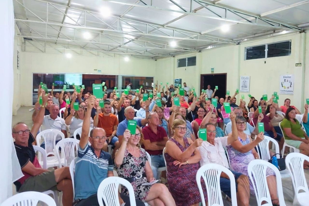 Um grande grupo de associados participa de uma assembleia da Sicredi Caminho das Águas. Sentados em cadeiras brancas, levantam cartões verdes com a palavra &quot;Assembleia&quot;, demonstrando engajamento e participação no evento cooperativo.