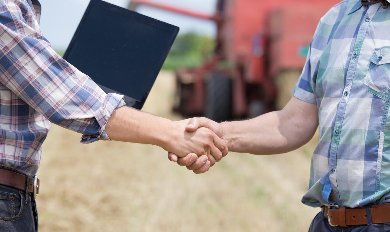Dois agricultores apertam as mãos em um campo, simbolizando parceria no agronegócio, com uma colheitadeira ao fundo.